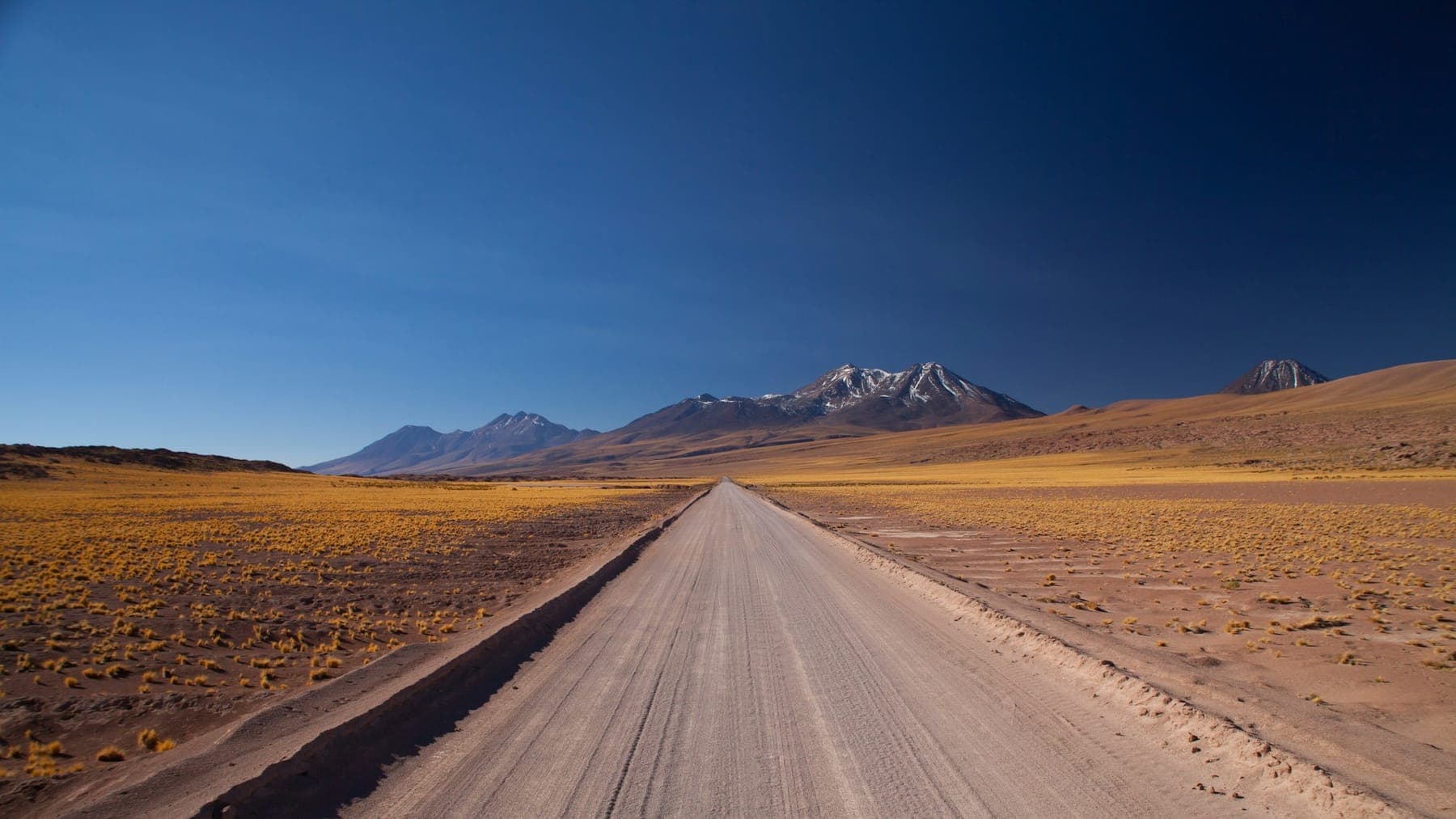 A dirt road running straight across the high desert toward snow-capped volcanoes near San Pedro de Atacama