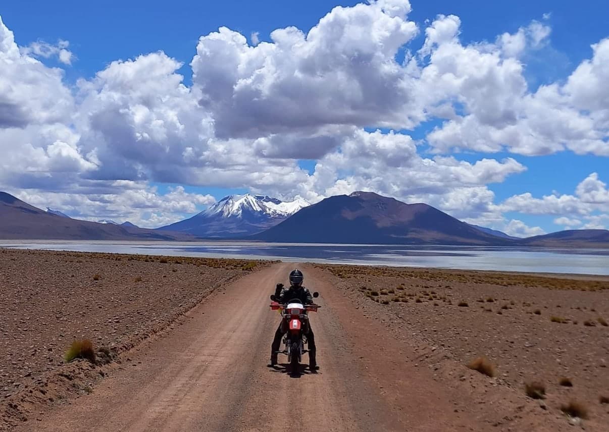 Ross on a red dual-sport bike on a dirt road across the Bolivian altiplano, with a salt lake and a snow-capped volcano behind
