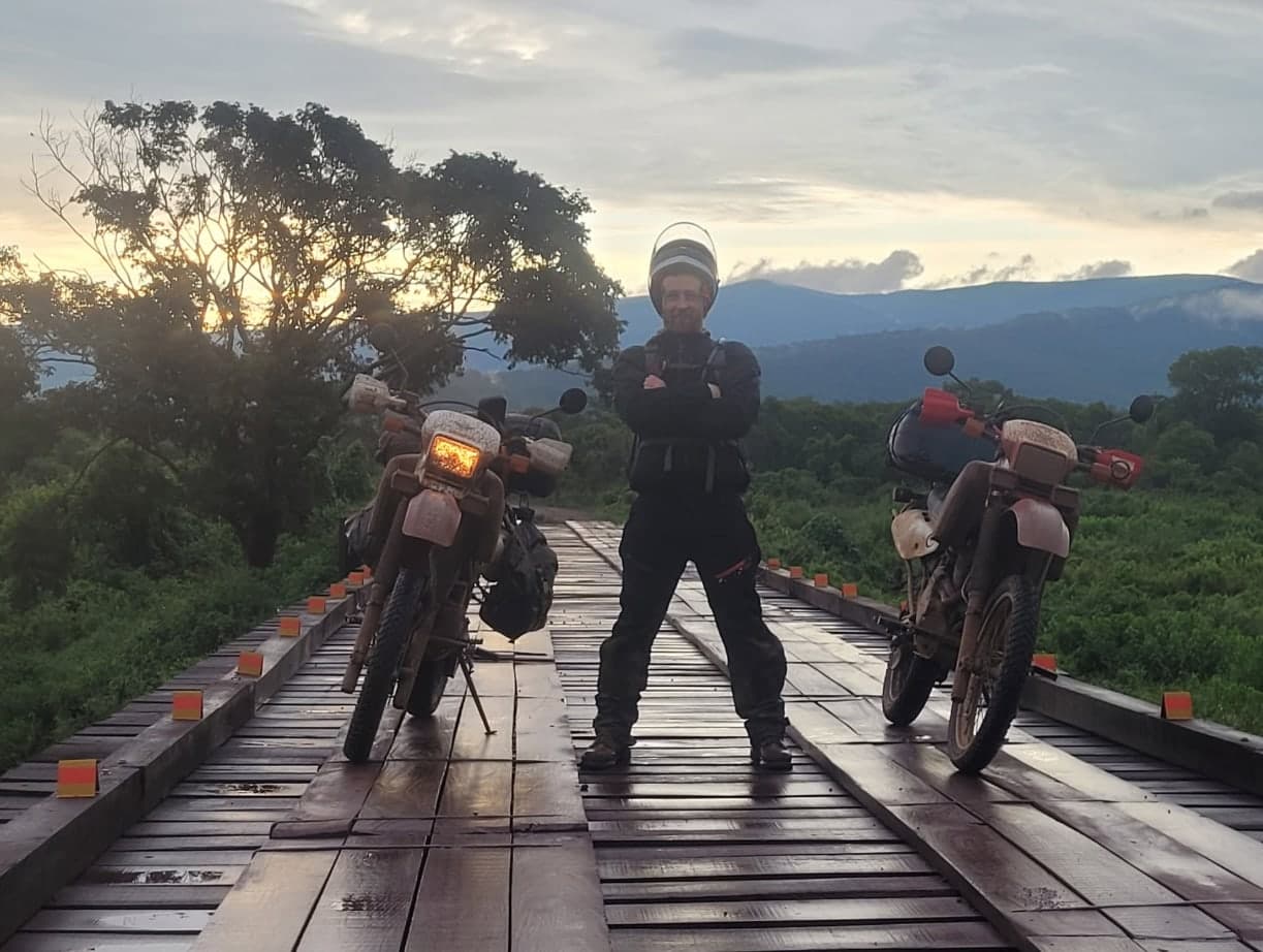 Ross in riding gear, arms crossed, between two mud-covered dual-sport bikes on a plank bridge at sunrise in the Pantanal near Corumbá, Brazil