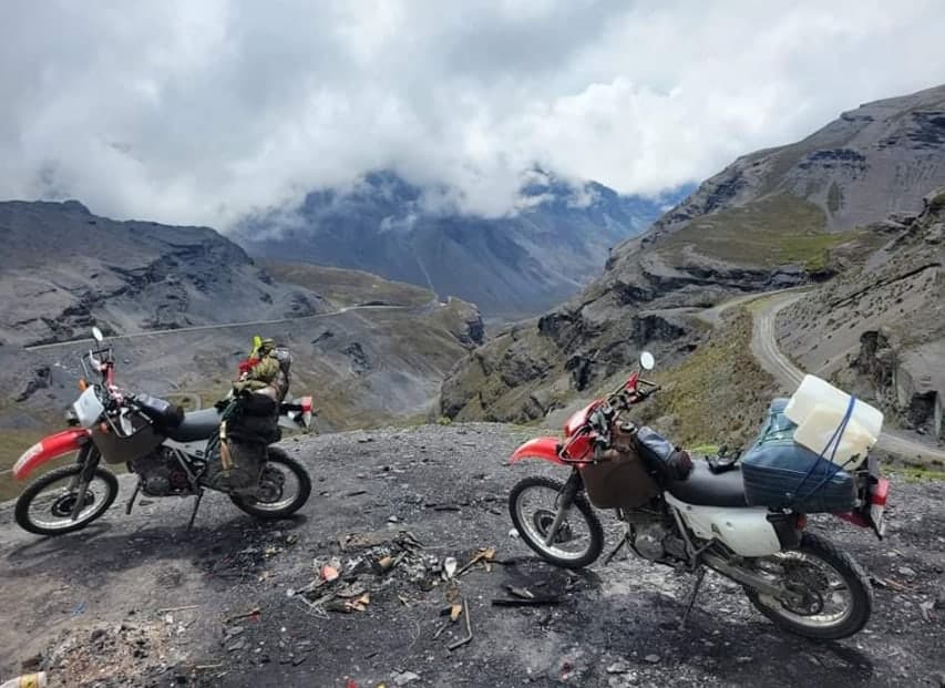 Two loaded dual-sport bikes parked on a high mountain pass with a dirt road winding below through the clouds