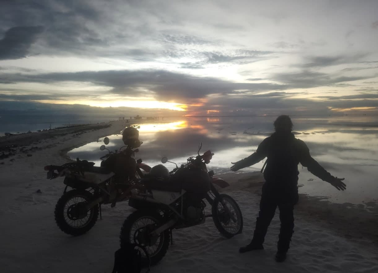 Two dual-sport bikes and Ross with arms spread at sunset on the flooded Salar de Uyuni
